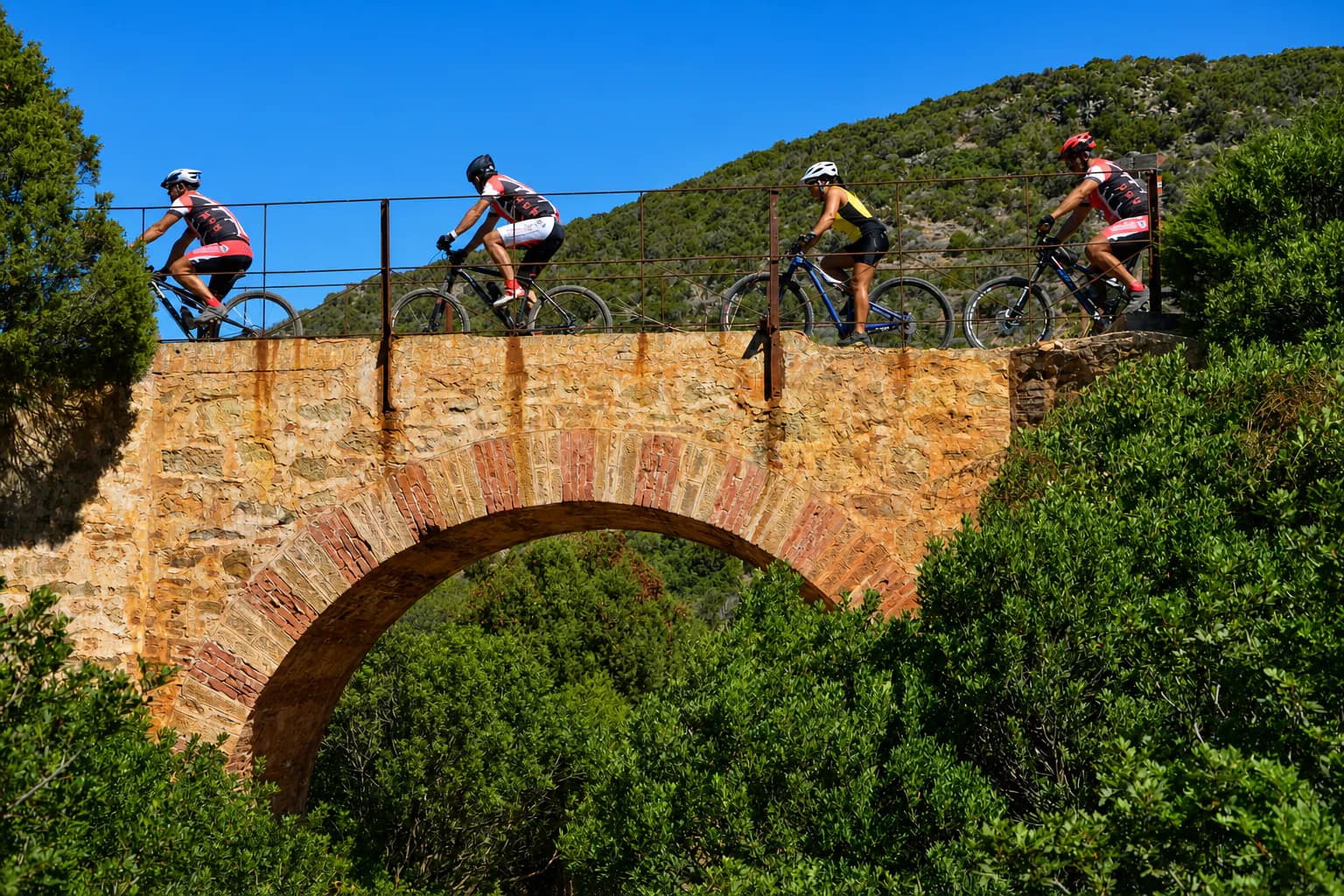 Four adult cyclists crossing a historic stone bridge on a Sardinian route